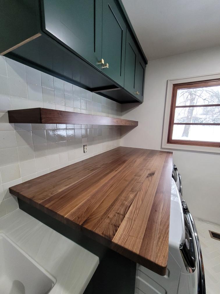 Dark green laundry room cabinets, walnut countertop, floating shelf under the cabinets.